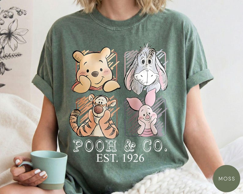 May include: Moss green t-shirt featuring four cartoon character portraits: Winnie the Pooh, Eeyore, Tigger, and Piglet. The words "POOH & CO. EST. 1926" are printed below the characters. The shirt has rolled-up sleeves.