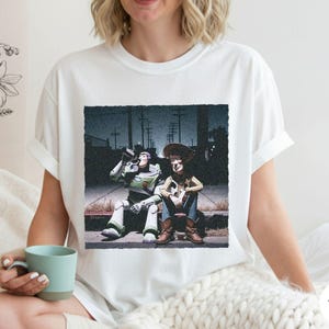 May include: A white short-sleeved t-shirt featuring a graphic print of two animated characters, a space ranger and a cowboy, sitting on a curb at night. The print shows the characters in a dark, urban setting with utility poles in the background.