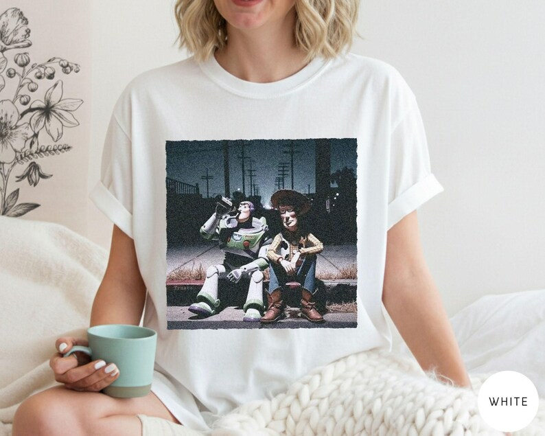 May include: A white short-sleeved t-shirt featuring a graphic print of two animated characters, a space ranger and a cowboy, sitting on a curb at night. The print shows the characters in a dark, urban setting with utility poles in the background.