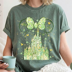 K&ouml;nnte beinhalten: Moosgr&uuml;nes T-Shirt mit einer skurrilen Illustration eines Schlosses, einer gro&szlig;en Schleife und Mickey Mouse-Silhouetten. Das Schloss ist in Gr&uuml;n- und Gelbt&ouml;nen gehalten, mit einem Aquarelleffekt. Die Schleife hat ein Tupfenmuster.