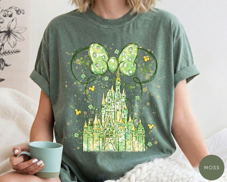 K&ouml;nnte beinhalten: Moosgr&uuml;nes T-Shirt mit einer skurrilen Illustration eines Schlosses, einer gro&szlig;en Schleife und Mickey Mouse-Silhouetten. Das Schloss ist in Gr&uuml;n- und Gelbt&ouml;nen gehalten, mit einem Aquarelleffekt. Die Schleife hat ein Tupfenmuster.