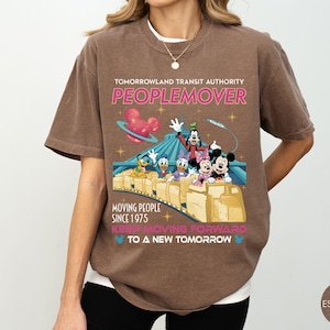 May include: Espresso-colored t-shirt featuring a graphic of Disney characters on a PeopleMover ride. The shirt includes the text "TOMORROWLAND TRANSIT AUTHORITY PEOPLEMOVER MOVING PEOPLE SINCE 1975 KEEP MOVING FORWARD TO A NEW TOMORROW."