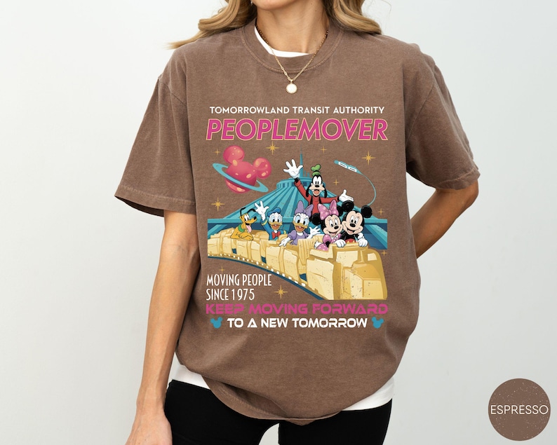 May include: Espresso-colored t-shirt featuring a graphic of Disney characters on a PeopleMover ride. The shirt includes the text "TOMORROWLAND TRANSIT AUTHORITY PEOPLEMOVER MOVING PEOPLE SINCE 1975 KEEP MOVING FORWARD TO A NEW TOMORROW."