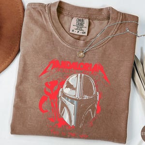 Op de afbeelding: Espresso-kleurig T-shirt met een rood en grijze afbeelding van een Mandalorian helm en tekst. Een zilveren ketting met een hanger ligt op het shirt. Het shirt heeft een ronde hals en een klein label bij de kraag.