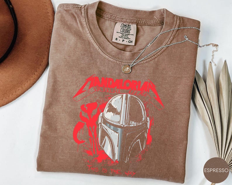 Op de afbeelding: Espresso-kleurig T-shirt met een rood en grijze afbeelding van een Mandalorian helm en tekst. Een zilveren ketting met een hanger ligt op het shirt. Het shirt heeft een ronde hals en een klein label bij de kraag.