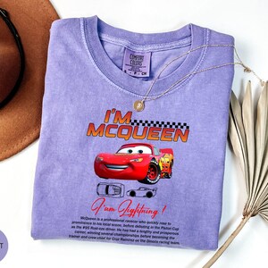 Puede incluir: Una camiseta violeta Comfort Colors con un gr&aacute;fico de Rayo McQueen de la pel&iacute;cula Cars. El dise&ntilde;o incluye el texto "I'M MCQUEEN" encima del coche y "I am Lightning!" debajo. Un collar dorado est&aacute; sobre la camiseta.