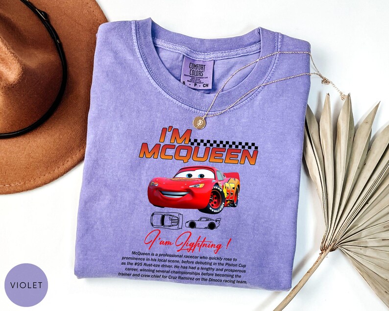 Puede incluir: Una camiseta violeta Comfort Colors con un gr&aacute;fico de Rayo McQueen de la pel&iacute;cula Cars. El dise&ntilde;o incluye el texto "I'M MCQUEEN" encima del coche y "I am Lightning!" debajo. Un collar dorado est&aacute; sobre la camiseta.