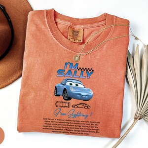 Puede incluir: Camiseta de color coral con un coche de dibujos animados azul con el texto "I'M SALLY" encima y "I am Lightning!" debajo. La camiseta tambi&eacute;n tiene una descripci&oacute;n del personaje del coche. Una cadena de oro y un sombrero marr&oacute;n tambi&eacute;n est&aacute;n en la imagen.