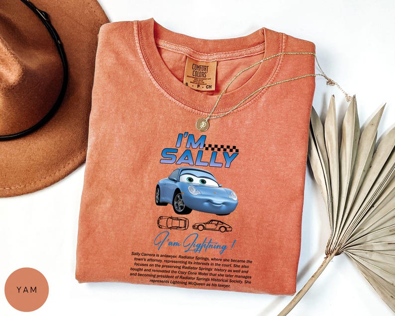 Puede incluir: Camiseta de color coral con un coche de dibujos animados azul con el texto "I'M SALLY" encima y "I am Lightning!" debajo. La camiseta tambi&eacute;n tiene una descripci&oacute;n del personaje del coche. Una cadena de oro y un sombrero marr&oacute;n tambi&eacute;n est&aacute;n en la imagen.