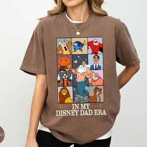 May include: Brown t-shirt with a graphic featuring Disney characters in a grid, including Mr. Incredible, Dory, and Scar. The text "IN MY DISNEY DAD ERA" is printed below the grid. The shirt is a relaxed fit.