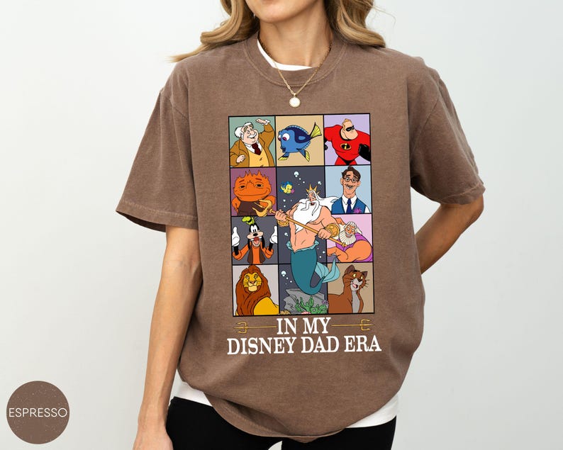 May include: Brown t-shirt with a graphic featuring Disney characters in a grid, including Mr. Incredible, Dory, and Scar. The text "IN MY DISNEY DAD ERA" is printed below the grid. The shirt is a relaxed fit.