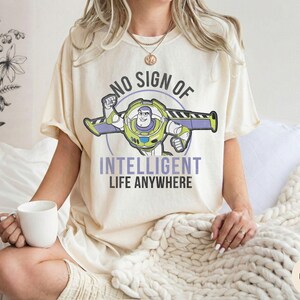 Puede incluir: Camiseta de color marfil con un dise&ntilde;o de Buzz Lightyear y el texto "NO SIGN OF INTELLIGENT LIFE ANYWHERE". El dise&ntilde;o incluye elementos verdes, morados y blancos. La camiseta es de corte holgado.