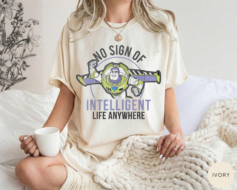 Puede incluir: Camiseta de color marfil con un dise&ntilde;o de Buzz Lightyear y el texto "NO SIGN OF INTELLIGENT LIFE ANYWHERE". El dise&ntilde;o incluye elementos verdes, morados y blancos. La camiseta es de corte holgado.