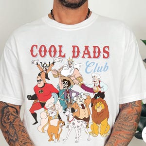 May include: White t-shirt with the text "COOL DADS Club" in red and blue. The shirt features cartoon characters including Mr. Incredible, Simba, and others. The shirt is a crew neck and has short sleeves.