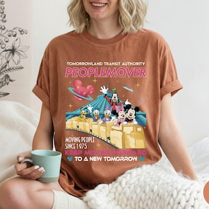 May include: Brown t-shirt featuring a colorful graphic of Disney characters on a PeopleMover ride. The text reads "Tomorrowland Transit Authority PEOPLEMOVER" and "Moving People Since 1975".