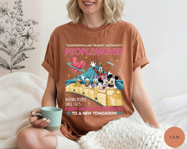 May include: Brown t-shirt featuring a colorful graphic of Disney characters on a PeopleMover ride. The text reads "Tomorrowland Transit Authority PEOPLEMOVER" and "Moving People Since 1975".