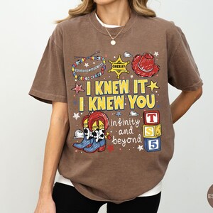 May include: A brown short-sleeve t-shirt featuring a graphic print with the text I KNEW IT I KNEW YOU, infinity and beyond, and T S 5. The design includes illustrations of a beaded bracelet, a sheriff badge, a red hat, cowboy boots, and blocks.