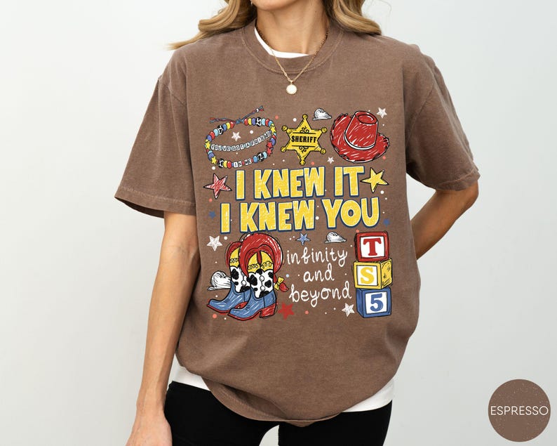 May include: A brown short-sleeve t-shirt featuring a graphic print with the text I KNEW IT I KNEW YOU, infinity and beyond, and T S 5. The design includes illustrations of a beaded bracelet, a sheriff badge, a red hat, cowboy boots, and blocks.