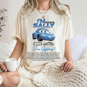 Puede incluir: Camiseta de color marfil con un coche de dibujos animados azul y el texto "I'M SALLY" encima y "I am Lightning!" debajo. La camiseta tambi&eacute;n incluye texto que describe al personaje Sally Carrera.