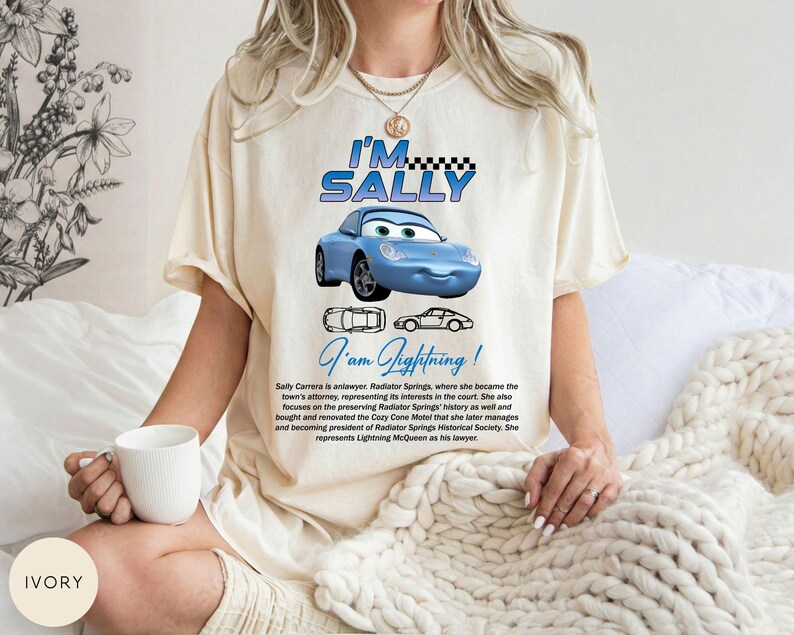 Puede incluir: Camiseta de color marfil con un coche de dibujos animados azul y el texto "I'M SALLY" encima y "I am Lightning!" debajo. La camiseta tambi&eacute;n incluye texto que describe al personaje Sally Carrera.