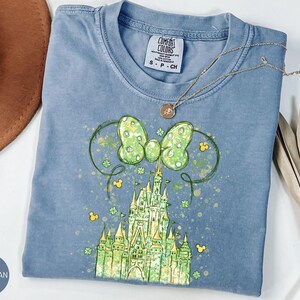 K&ouml;nnte beinhalten: Ein blaues Jeans-T-Shirt mit einer gr&uuml;nen und gelben Schlossgrafik. Die Grafik enth&auml;lt eine gr&uuml;ne Schleife mit Punkten, Mickey Mouse Ohren und Kleebl&auml;ttern. Eine goldene Halskette mit Anh&auml;nger liegt auf dem Shirt.