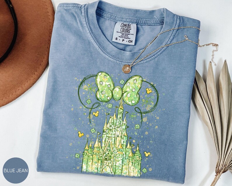 K&ouml;nnte beinhalten: Ein blaues Jeans-T-Shirt mit einer gr&uuml;nen und gelben Schlossgrafik. Die Grafik enth&auml;lt eine gr&uuml;ne Schleife mit Punkten, Mickey Mouse Ohren und Kleebl&auml;ttern. Eine goldene Halskette mit Anh&auml;nger liegt auf dem Shirt.