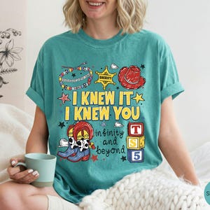 May include: A seafoam green t-shirt featuring a graphic print with the text I KNEW IT I KNEW YOU and infinity and beyond. The design includes a colorful beaded bracelet, a yellow sheriff star, a red cowboy hat, cow print boots, and blocks.