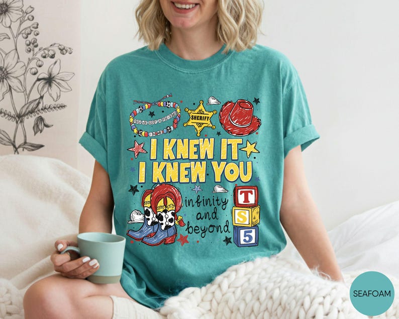 May include: A seafoam green t-shirt featuring a graphic print with the text I KNEW IT I KNEW YOU and infinity and beyond. The design includes a colorful beaded bracelet, a yellow sheriff star, a red cowboy hat, cow print boots, and blocks.