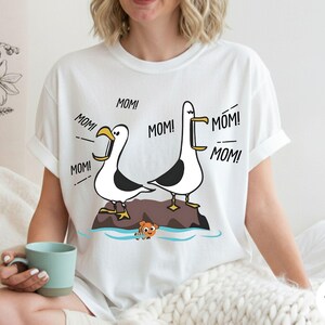 May include: White t-shirt with a cartoon design of two seagulls on a rock, with the word "MOM!" coming from their beaks. An orange fish is also in the image. The shirt is a casual, unisex style.