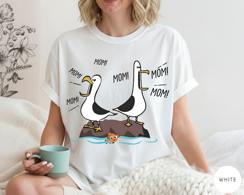 May include: White t-shirt with a cartoon design of two seagulls on a rock, with the word "MOM!" coming from their beaks. An orange fish is also in the image. The shirt is a casual, unisex style.