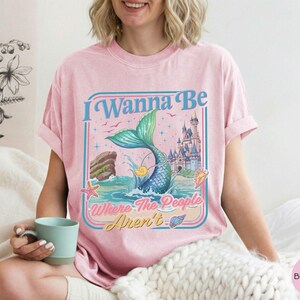May include: Pink t-shirt with a graphic featuring a mermaid tail, castle, and the text "I Wanna Be Where The People Aren't." The design includes a mermaid tail, waves, and seashells. The shirt is a light pink color.