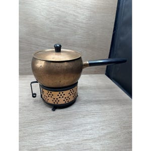 Puede incluir: Una olla de fondue de cobre vintage con asa y tapa negras. La olla se asienta sobre un soporte de metal negro con un diseño perforado. La olla de fondue es una pieza clásica para el entretenimiento.