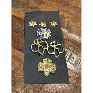 May include: A collection of Girl Scout pins and badges on a black card. The pins include gold stars, a blue and gold emblem, two gold girl figure pins, and a gold trefoil pin.