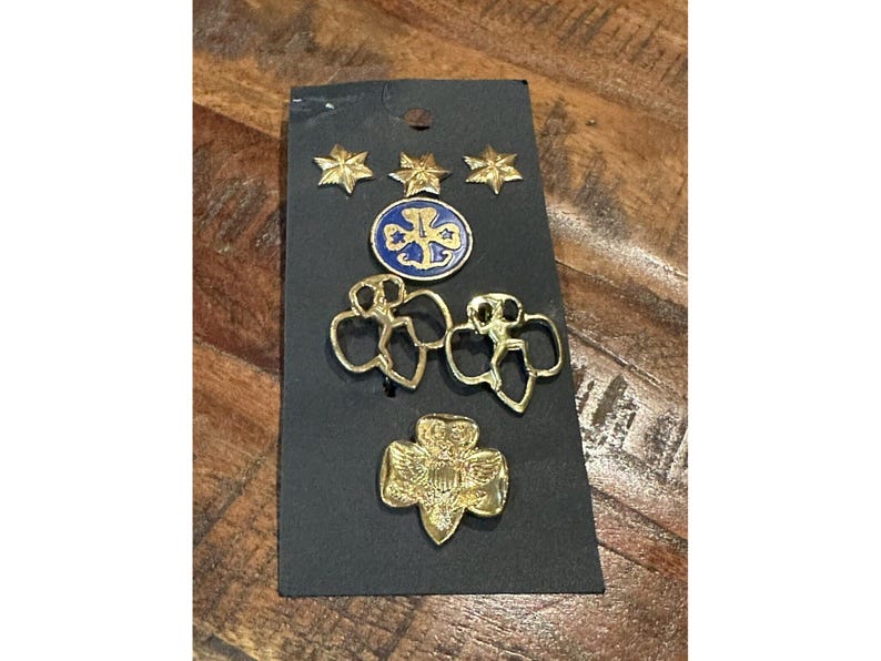 May include: A collection of Girl Scout pins and badges on a black card. The pins include gold stars, a blue and gold emblem, two gold girl figure pins, and a gold trefoil pin.