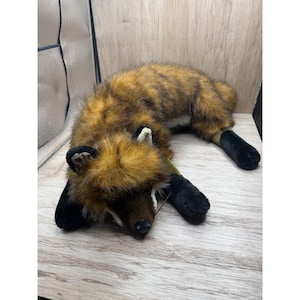 May include: A soft toy fox with brown and black fur, black paws, and white details. The toy is lying on a wooden surface. The fox has a realistic design and is suitable for children.