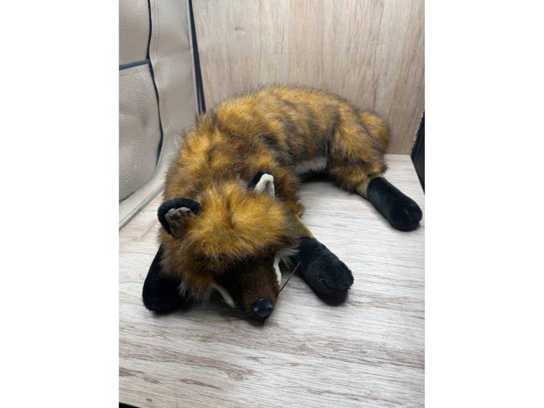 May include: A soft toy fox with brown and black fur, black paws, and white details. The toy is lying on a wooden surface. The fox has a realistic design and is suitable for children.