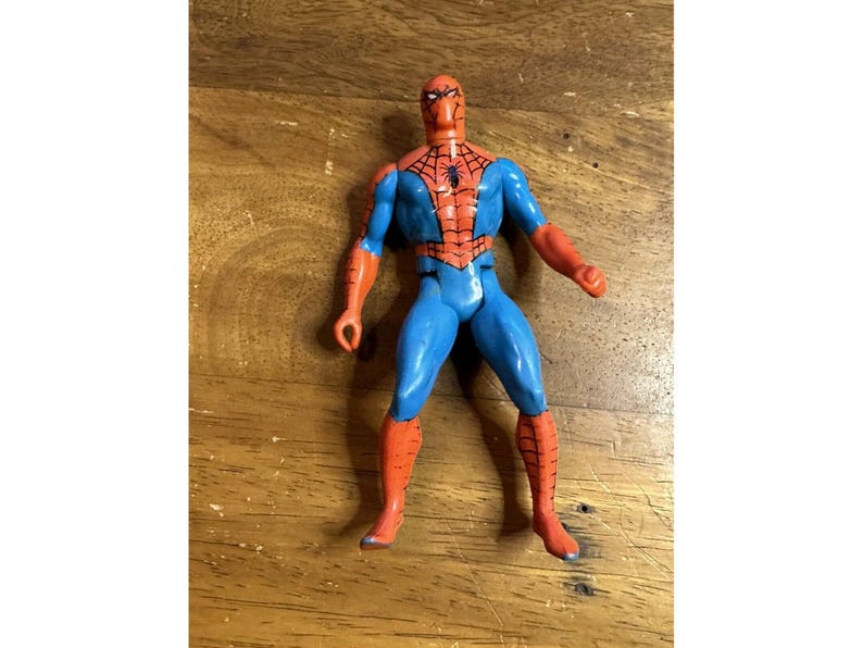 Puede incluir: Una figura de acci&oacute;n de pl&aacute;stico de un superh&eacute;roe con un traje rojo y azul que presenta un emblema de ara&ntilde;a negro en el pecho. La figura tiene una m&aacute;scara roja con parches oculares blancos, botas y guantes rojos, y torso y piernas de color azul.