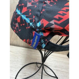 May include: A baseball cap featuring a black base with a vibrant geometric pattern in red, orange, and light blue. A small blue fabric tag with the yellow text PENDLETON is stitched onto the side of the cap, which is displayed on a black stand.