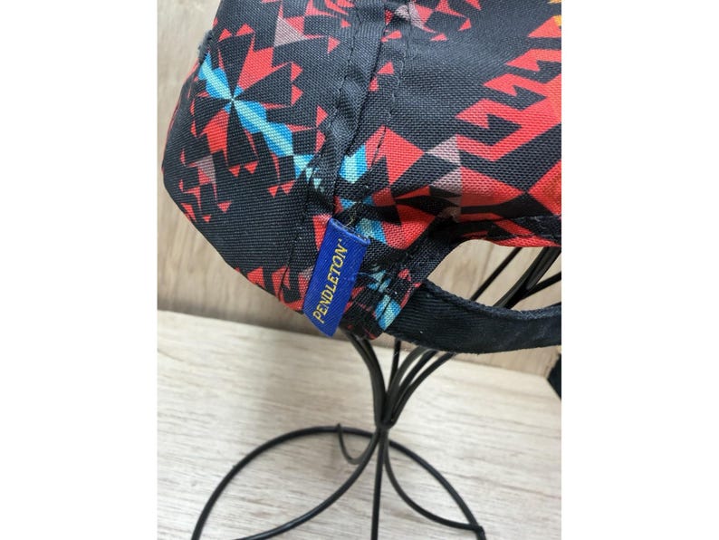 May include: A baseball cap featuring a black base with a vibrant geometric pattern in red, orange, and light blue. A small blue fabric tag with the yellow text PENDLETON is stitched onto the side of the cap, which is displayed on a black stand.