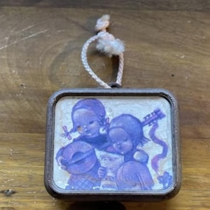 Puede incluir: Caja de música vintage con marco rectangular e ilustración detallada de dos niños tocando instrumentos. La imagen es en tonos morados y blancos, con una cuerda decorativa y borla en la parte superior.