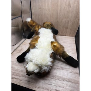 May include: A soft toy fox, lying on its back, with a white chest, brown and orange fur, and black paws. The toy is placed on a light-coloured wooden surface, highlighting its soft texture and playful design.