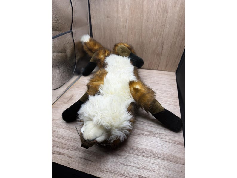May include: A soft toy fox, lying on its back, with a white chest, brown and orange fur, and black paws. The toy is placed on a light-coloured wooden surface, highlighting its soft texture and playful design.
