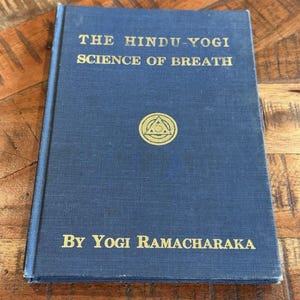 Puede incluir: Un libro de tapa dura azul titulado The Hindu-Yogi Science of Breath por Yogi Ramacharaka. La portada presenta texto en relieve dorado y un emblema circular central con un triángulo y rayos de sol, sobre un acabado de tela texturizada.