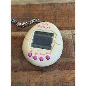 May include: A vintage Tamagotchi Puppy digital pet, egg-shaped and cream-colored, with a small screen and pink buttons. The text "Ta-Ta Puppy" is visible. It is attached to a silver chain.