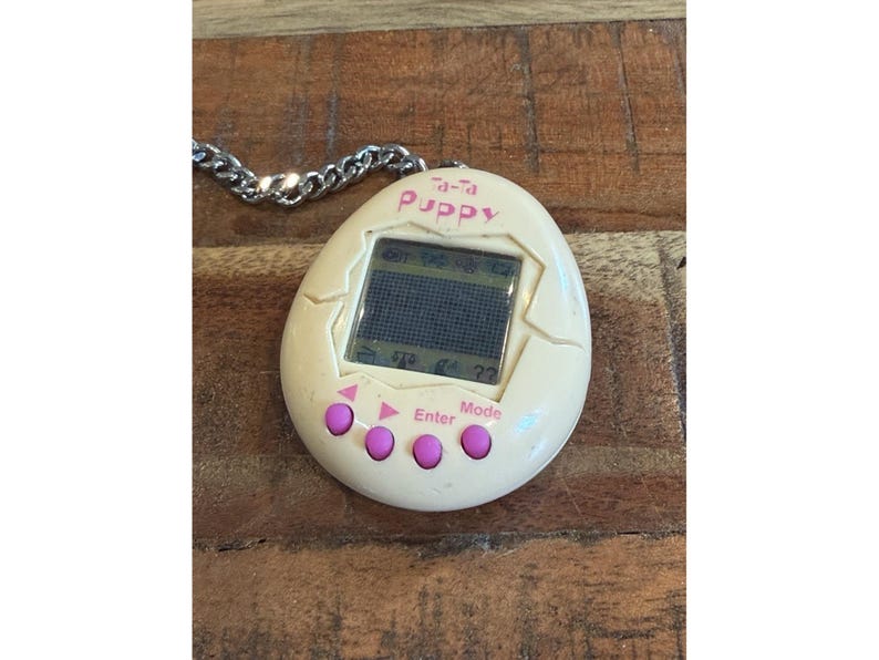 May include: A vintage Tamagotchi Puppy digital pet, egg-shaped and cream-colored, with a small screen and pink buttons. The text "Ta-Ta Puppy" is visible. It is attached to a silver chain.