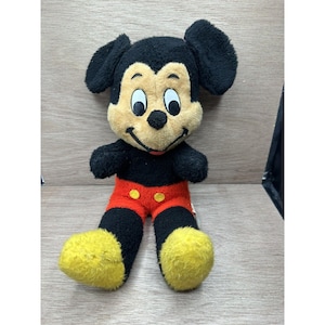 May include: A vintage Mickey Mouse plush toy. The plush features a black body, ears, and legs, with a tan face and yellow feet. Mickey is wearing red shorts with yellow buttons. The toy is sitting on a wooden surface.