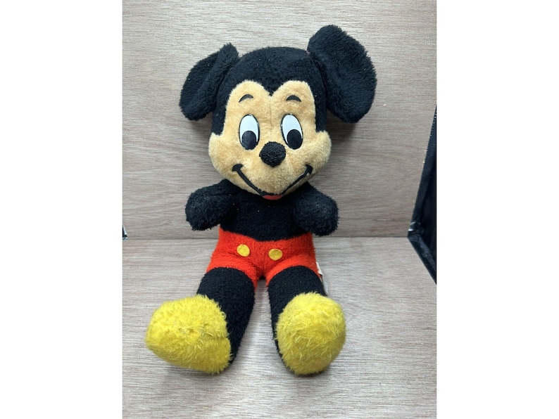 May include: A vintage Mickey Mouse plush toy. The plush features a black body, ears, and legs, with a tan face and yellow feet. Mickey is wearing red shorts with yellow buttons. The toy is sitting on a wooden surface.