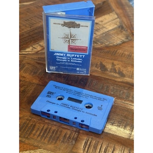 Pode incluir: Uma fita cassete azul e sua caixa com o álbum "Changes in Latitudes, Changes in Attitudes" de Jimmy Buffett. A fita cassete é azul com texto branco. A caixa é transparente com um rótulo azul.