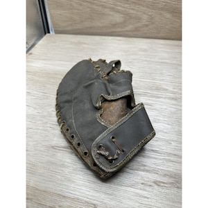 Peut inclure: Un gant de baseball ancien en cuir marron foncé. Le gant présente une apparence usée avec des coutures visibles et une sangle de poignet. Il est conçu pour attraper une balle de baseball et date probablement du début ou du milieu du XXe siècle.