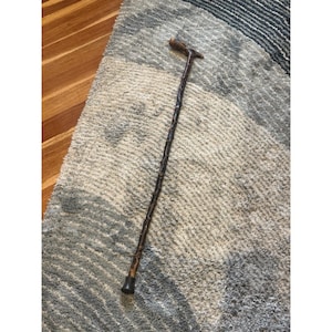 May include: A dark brown wooden walking cane with a curved handle and a black rubber tip. The cane has a textured, natural wood grain appearance. The cane is resting on a gray and white patterned rug.